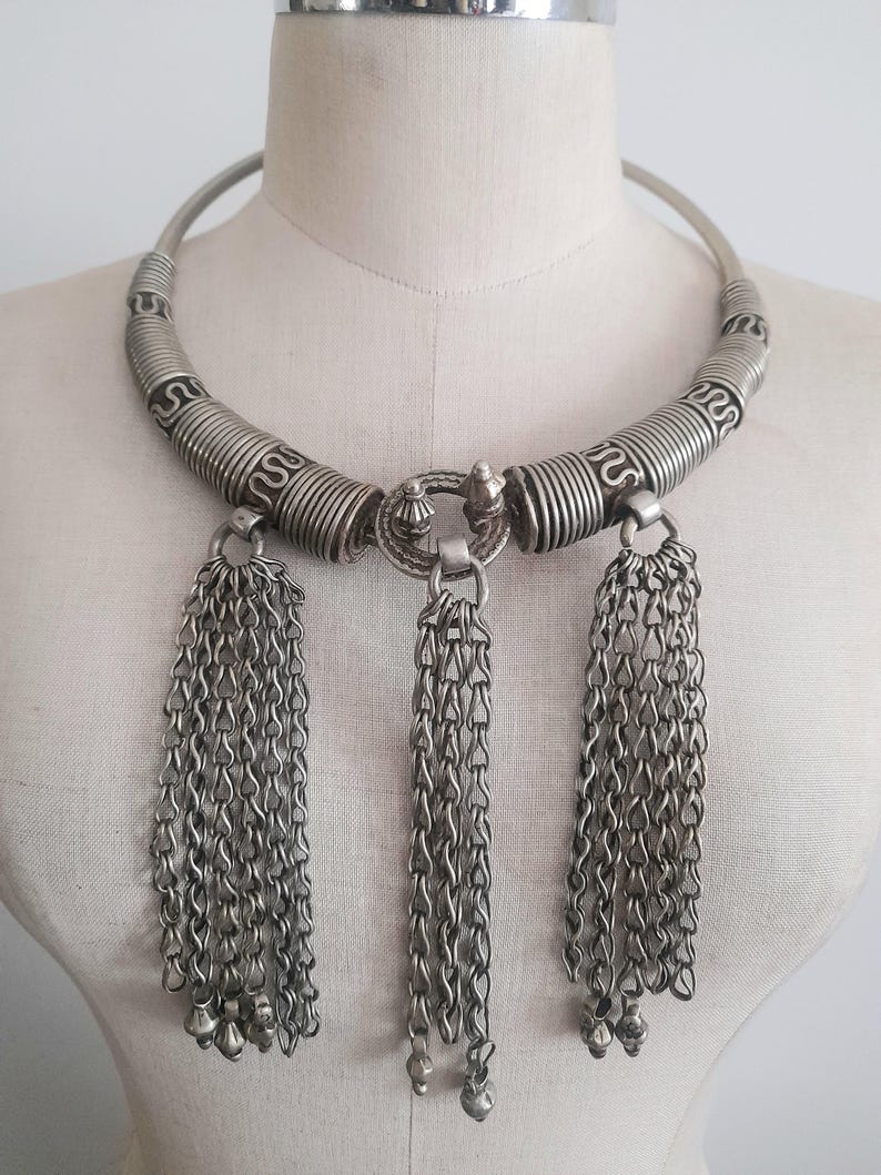 May include: Silver-toned statement necklace with a rigid collar and three chain tassel pendants. The collar features spiral and linear detailing. The tassels are composed of delicate chain links and small bell-shaped charms.