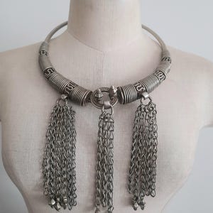 May include: Silver-toned statement necklace with a rigid collar and three chain tassel pendants. The collar features coiled and patterned sections. Each tassel ends with small, bell-shaped charms. The necklace is displayed on a mannequin.