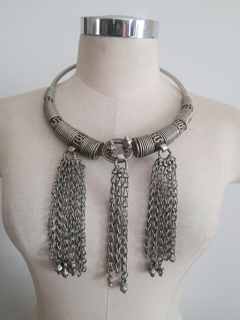 May include: Silver-toned statement necklace with a rigid collar and three chain tassel pendants. The collar features coiled and patterned sections. Each tassel ends with small, bell-shaped charms. The necklace is displayed on a mannequin.
