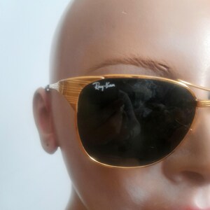 May include: Gold-framed Ray-Ban sunglasses with dark lenses. The sunglasses have a classic aviator shape with a double bridge and the Ray-Ban logo on the lens. The frame has a textured design.