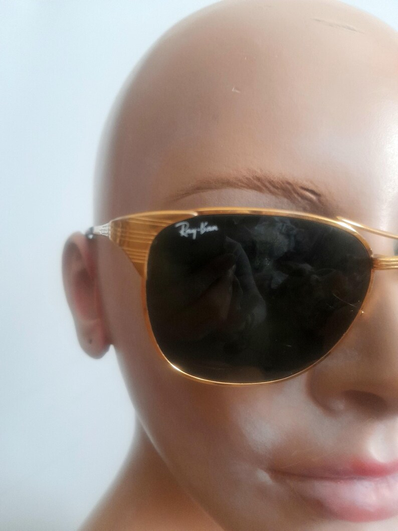 May include: Gold-framed Ray-Ban sunglasses with dark lenses. The sunglasses have a classic aviator shape with a double bridge and the Ray-Ban logo on the lens. The frame has a textured design.