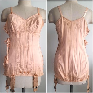 Pode incluir: Um espartilho vintage cor de pêssego com decote em V, alças delicadas e detalhes de botões. O espartilho apresenta costuras verticais, cadarços laterais e ligas presas. A peça de roupa é exibida em um manequim.