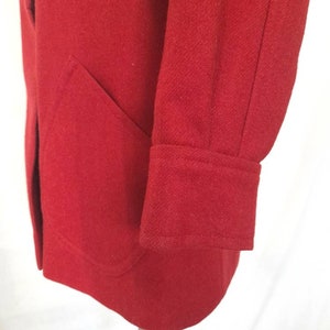 K&ouml;nnte beinhalten: Ein roter Wollmantel mit einer einzigen Tasche auf der linken Seite und einem Knopfverschluss. Der Mantel hat einen langen &Auml;rmel mit einer gefalteten Manschette.