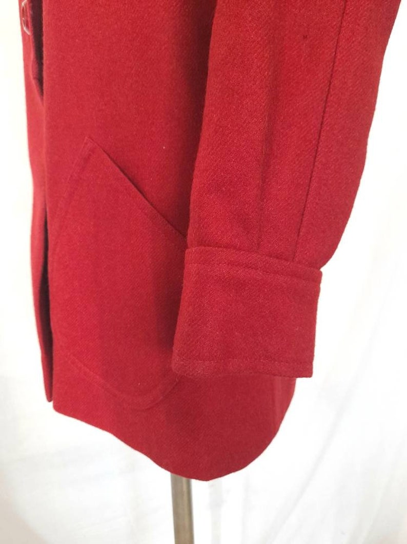 K&ouml;nnte beinhalten: Ein roter Wollmantel mit einer einzigen Tasche auf der linken Seite und einem Knopfverschluss. Der Mantel hat einen langen &Auml;rmel mit einer gefalteten Manschette.