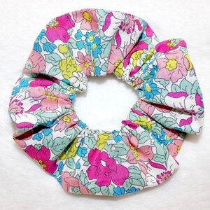May include: A floral scrunchie featuring a pattern of pink, blue, and yellow flowers on a white background. The fabric is gathered and elasticized, creating a ruffled effect. This hair accessory is suitable for various hairstyles.