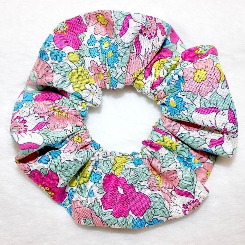 May include: A floral scrunchie featuring a pattern of pink, blue, and yellow flowers on a white background. The fabric is gathered and elasticized, creating a ruffled effect. This hair accessory is suitable for various hairstyles.