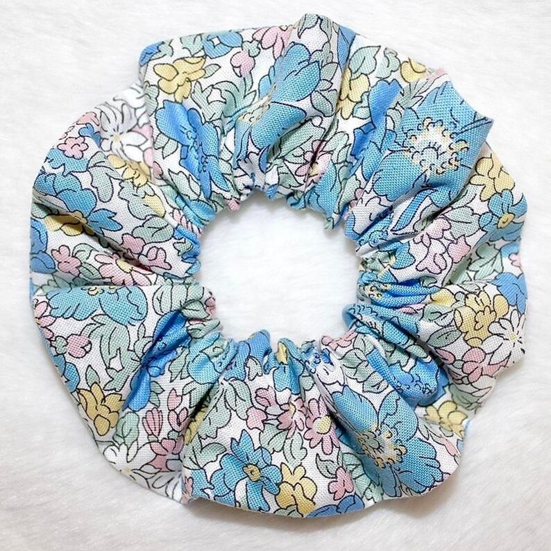 May include: A floral scrunchie featuring a pattern of blue, pink, and yellow flowers on a white background. The fabric is gathered around an elastic band, creating a ruffled effect. This hair accessory is designed for tying up hair.