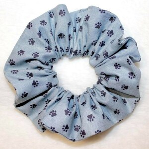 May include: A light blue scrunchie with a repeating pattern of dark gray paw prints. The fabric is gathered around an elastic band, creating a ruffled appearance. This hair accessory is suitable for various hairstyles.