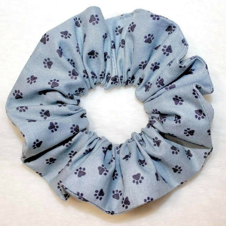 May include: A light blue scrunchie with a repeating pattern of dark gray paw prints. The fabric is gathered around an elastic band, creating a ruffled appearance. This hair accessory is suitable for various hairstyles.