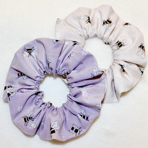May include: Two scrunchies with bee patterns. One scrunchie is lavender with white and black bees, and the other is light beige with black and yellow bees. Both scrunchies are made of fabric.