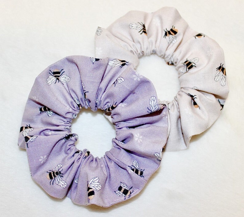 May include: Two scrunchies with bee patterns. One scrunchie is lavender with white and black bees, and the other is light beige with black and yellow bees. Both scrunchies are made of fabric.