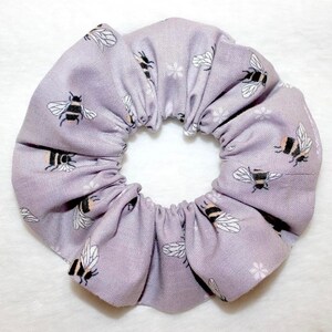 May include: A lavender scrunchie with a repeating pattern of black and yellow bees and small white floral accents. The fabric is gathered, creating a textured, circular shape. This hair accessory is perfect for adding a touch of whimsy to any hairstyle.