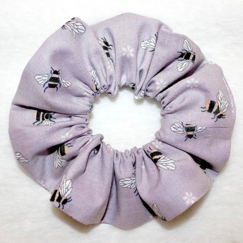May include: A lavender scrunchie with a repeating pattern of black and yellow bees and small white floral accents. The fabric is gathered, creating a textured, circular shape. This hair accessory is perfect for adding a touch of whimsy to any hairstyle.