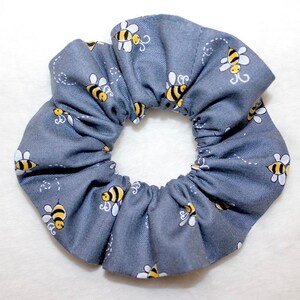May include: A gray scrunchie with a repeating pattern of yellow and black bees. The bees are stylized with white wings and are scattered across the fabric. The scrunchie is made of a soft, gathered material.