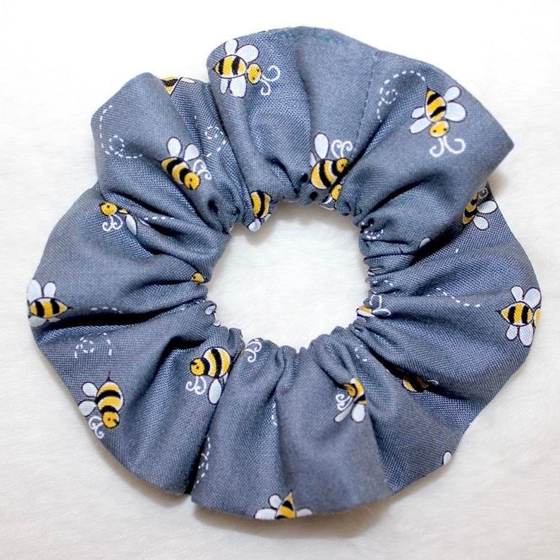 May include: A gray scrunchie with a repeating pattern of yellow and black bees. The bees are stylized with white wings and are scattered across the fabric. The scrunchie is made of a soft, gathered material.