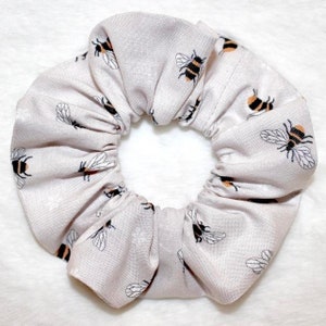 May include: A light gray scrunchie with a repeating pattern of black and yellow bees. The scrunchie is made of fabric and is gathered to create a ruffled effect. The background is white.
