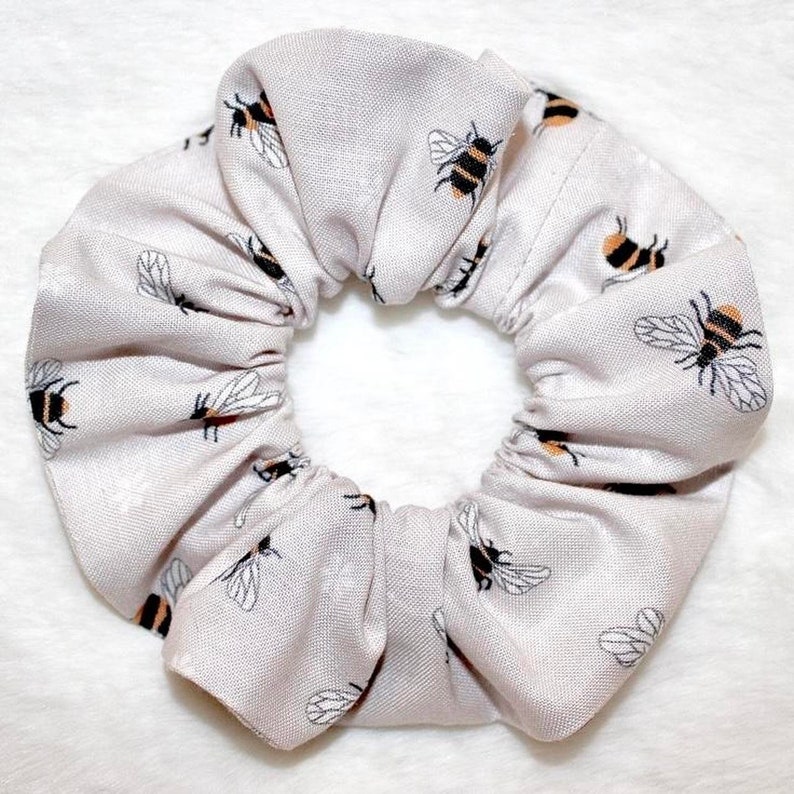 May include: A light gray scrunchie with a repeating pattern of black and yellow bees. The scrunchie is made of fabric and is gathered to create a ruffled effect. The background is white.