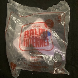 May include: A purple plastic toy from the Disney movie Ralph Breaks the Internet. The toy is a character named  "Vanellope Von Schweetz" from the movie. The toy is packaged in a clear plastic bag with a white label that says "Ralph Breaks the Internet" and "Disney".