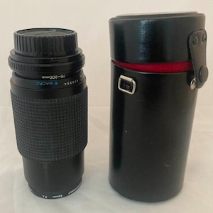 May include: A black camera lens with a silver ring and the text "75-200mm" and "MACRO" printed on it. The lens is in a black leather case with a red lining.