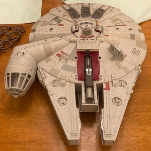 Puede incluir: Una nave espacial de juguete Millennium Falcon blanca y gris con detalles rojos y negros. La nave espacial tiene una cabina, un motor grande y una cápsula de escape desmontable.