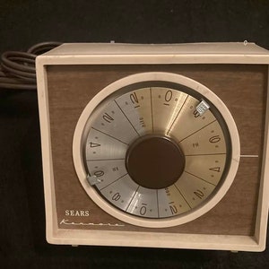 May include: A vintage Sears Kenmore timer with a white face and brown woodgrain trim. The timer has a round dial with black numbers and a knob in the center. The dial is set to 12:00.