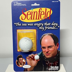 May include: A Seinfeld-themed collectible featuring a Titleist golf ball. The packaging is blue with the show's logo and a quote. The image of George Costanza holding a golf ball is also on the packaging.