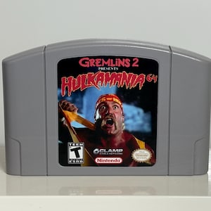 May include: A gray Nintendo 64 video game cartridge for "Gremlins 2 Presents Hulkamania 64." The label features a colorful image of Hulk Hogan and game title text. The game is rated "T" for Teen.