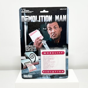 May include: A plastic action figure packaging with a photo of Sylvester Stallone as John Spartan, the Demolition Man, holding a stack of morality violation slips. The packaging is red, white, and black with the text "Demolition Man" in large white letters. The text "Morality Violation Slips (8 ct.)" is printed on the packaging. The packaging also includes a smaller card with the text "Morality Violation" and details about the character's morality violation.