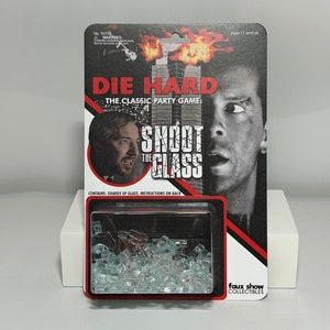 May include: A Die Hard themed party game called "Shoot the Glass". The game includes a box with shards of glass inside. The box has a picture of Bruce Willis as John McClane from the movie Die Hard. The box also has the text "Die Hard: The Classic Party Game" and "Shoot the Glass".