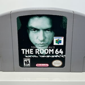 K&ouml;nnte beinhalten: Eine graue Nintendo 64-Spielkassette mit einem Schwarzwei&szlig;bild von Tommy Wiseau auf der Vorderseite. Der Spieltitel lautet "Tommy Wiseau's The Room 64" wobei der Text "The Room" in einer gr&ouml;&szlig;eren Schriftart dargestellt ist. Das Spiel ist von der ESRB mit "M" f&uuml;r Mature bewertet.