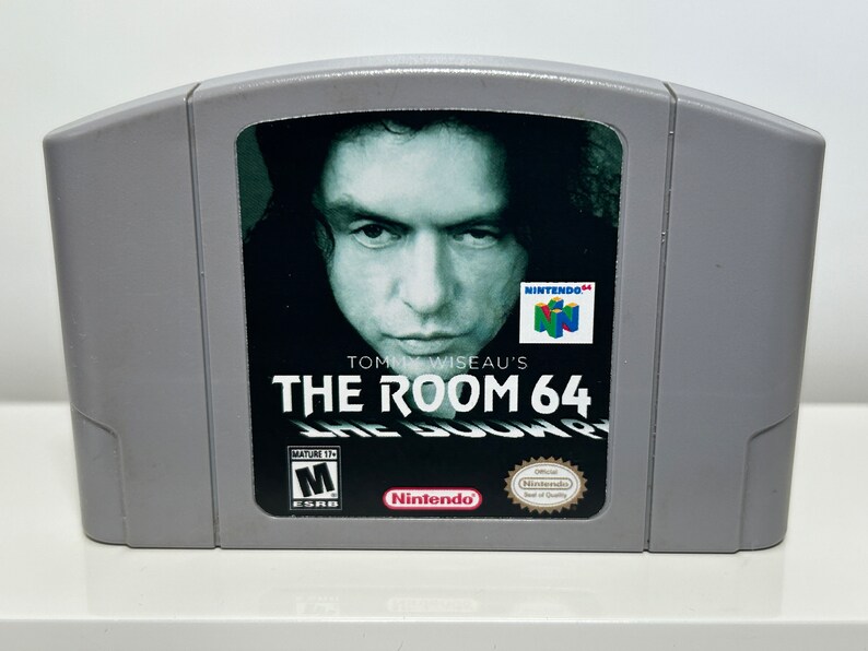 K&ouml;nnte beinhalten: Eine graue Nintendo 64-Spielkassette mit einem Schwarzwei&szlig;bild von Tommy Wiseau auf der Vorderseite. Der Spieltitel lautet "Tommy Wiseau's The Room 64" wobei der Text "The Room" in einer gr&ouml;&szlig;eren Schriftart dargestellt ist. Das Spiel ist von der ESRB mit "M" f&uuml;r Mature bewertet.