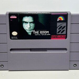 K&ouml;nnte beinhalten: Super Nintendo Entertainment System-Spielkassette f&uuml;r "Super Tommy Wiseau's The Room". Die Kassette ist grau mit einem schwarzen Etikett, das ein Portr&auml;t von Tommy Wiseau und den Spieltitel zeigt.