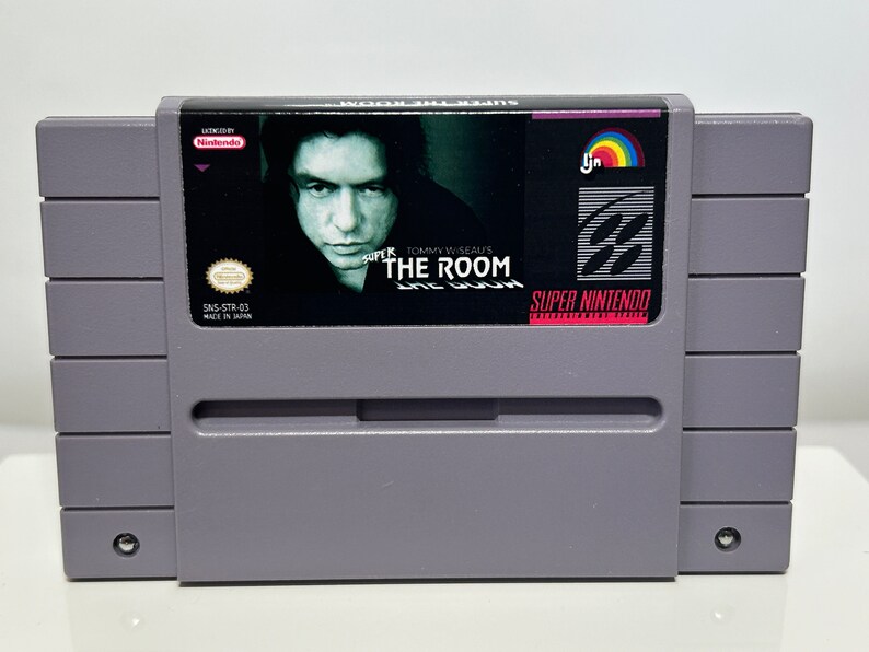 K&ouml;nnte beinhalten: Super Nintendo Entertainment System-Spielkassette f&uuml;r "Super Tommy Wiseau's The Room". Die Kassette ist grau mit einem schwarzen Etikett, das ein Portr&auml;t von Tommy Wiseau und den Spieltitel zeigt.