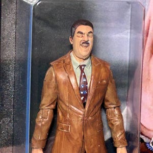 Pu&ograve; includere: Una action figure marrone di un uomo in giacca e cravatta e camicia. La figura &egrave; in una custodia di plastica trasparente. L'abito &egrave; di colore marrone medio e la cravatta ha un motivo a pois rossi e blu.