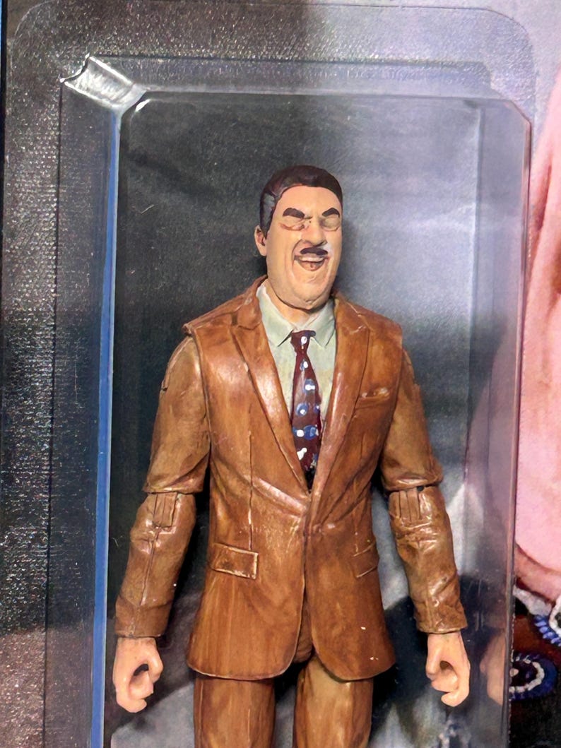 Pu&ograve; includere: Una action figure marrone di un uomo in giacca e cravatta e camicia. La figura &egrave; in una custodia di plastica trasparente. L'abito &egrave; di colore marrone medio e la cravatta ha un motivo a pois rossi e blu.