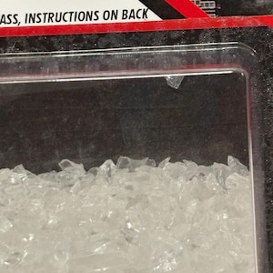 K&ouml;nnte beinhalten: Eine durchsichtige Kunststoff-Blisterverpackung mit kleinen, gezackten, transparenten Glasfragmenten. Die Verpackung hat einen rot-schwarzen Rand mit dem Text "ASS, INSTRUCTIONS ON BACK", der in Schwarz auf wei&szlig;em Grund gedruckt ist.