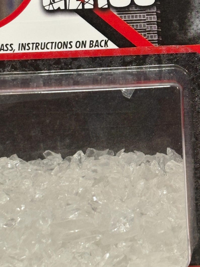 K&ouml;nnte beinhalten: Eine durchsichtige Kunststoff-Blisterverpackung mit kleinen, gezackten, transparenten Glasfragmenten. Die Verpackung hat einen rot-schwarzen Rand mit dem Text "ASS, INSTRUCTIONS ON BACK", der in Schwarz auf wei&szlig;em Grund gedruckt ist.