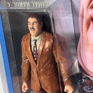 Pu&ograve; includere: Action figure marrone di un uomo in abito marrone, camicia bianca e cravatta fantasia. La figura &egrave; in una confezione di plastica trasparente con il nome "C. MAC" visibile. La figura ha un'espressione sorridente.