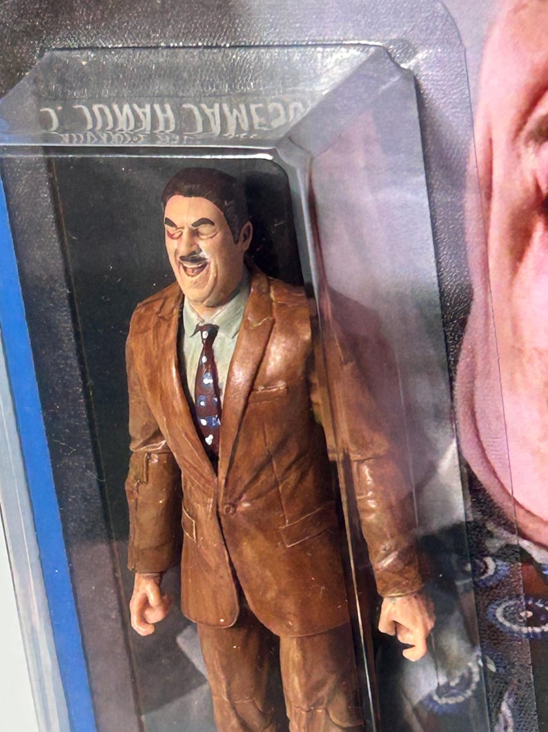 Pu&ograve; includere: Action figure marrone di un uomo in abito marrone, camicia bianca e cravatta fantasia. La figura &egrave; in una confezione di plastica trasparente con il nome "C. MAC" visibile. La figura ha un'espressione sorridente.