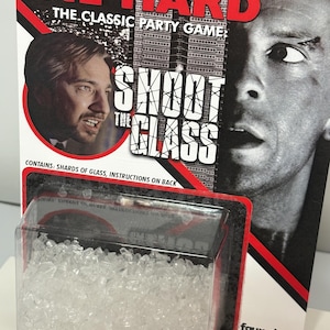 K&ouml;nnte beinhalten: Ein Die Hard The Classic Party Game Shoot The Glass Spielset in einer Blisterverpackung mit durchsichtigen Glassplittern. Die Verpackung zeigt den Filmtitel, ein M&auml;nnerportr&auml;t, einen Wolkenkratzer und den Text Contains: Shards of glass.
