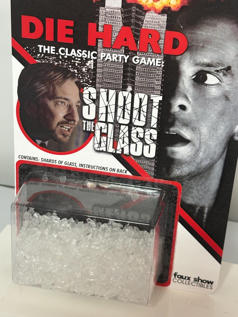 K&ouml;nnte beinhalten: Ein Die Hard The Classic Party Game Shoot The Glass Spielset in einer Blisterverpackung mit durchsichtigen Glassplittern. Die Verpackung zeigt den Filmtitel, ein M&auml;nnerportr&auml;t, einen Wolkenkratzer und den Text Contains: Shards of glass.