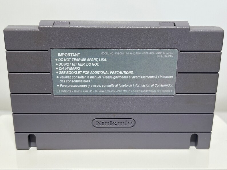 K&ouml;nnte beinhalten: Eine graue Super Nintendo Entertainment System (SNES) Spielkassette mit dem Text "IMPORTANT" und einer Liste von Warnhinweisen. Die Kassette tr&auml;gt das Nintendo-Logo unten.