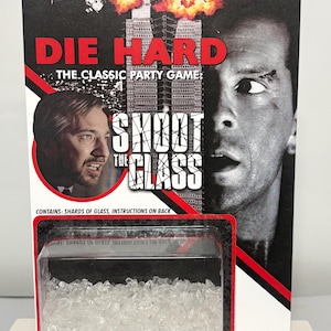 K&ouml;nnte beinhalten: Ein Die Hard Shoot The Glass Partyspiel in Original-Blisterverpackung. Die Karte zeigt Filmmotive, den Titel in roter und wei&szlig;er Schrift und eine klare Kunststoffblase am unteren Rand mit kleinen, durchsichtigen, zackigen Glassplittern.