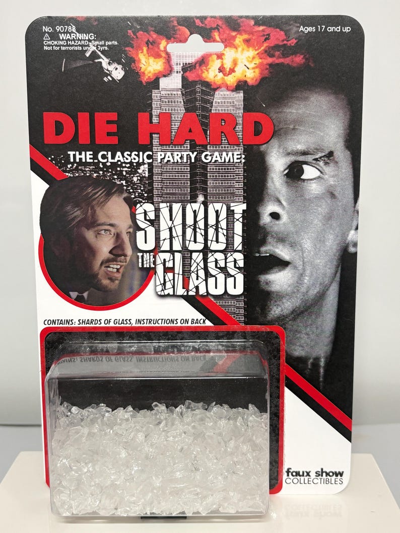 K&ouml;nnte beinhalten: Ein Die Hard Shoot The Glass Partyspiel in Original-Blisterverpackung. Die Karte zeigt Filmmotive, den Titel in roter und wei&szlig;er Schrift und eine klare Kunststoffblase am unteren Rand mit kleinen, durchsichtigen, zackigen Glassplittern.