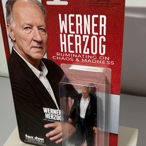 K&ouml;nnte beinhalten: Sammelfigur von Werner Herzog in einer transparenten Kunststoffverpackung. Die Verpackung zeigt ein Foto von Herzog und den Text "WERNER HERZOG RUMINATING ON CHAOS & MADNESS". Die Figur tr&auml;gt einen schwarzen Anzug und ein wei&szlig;es Hemd.