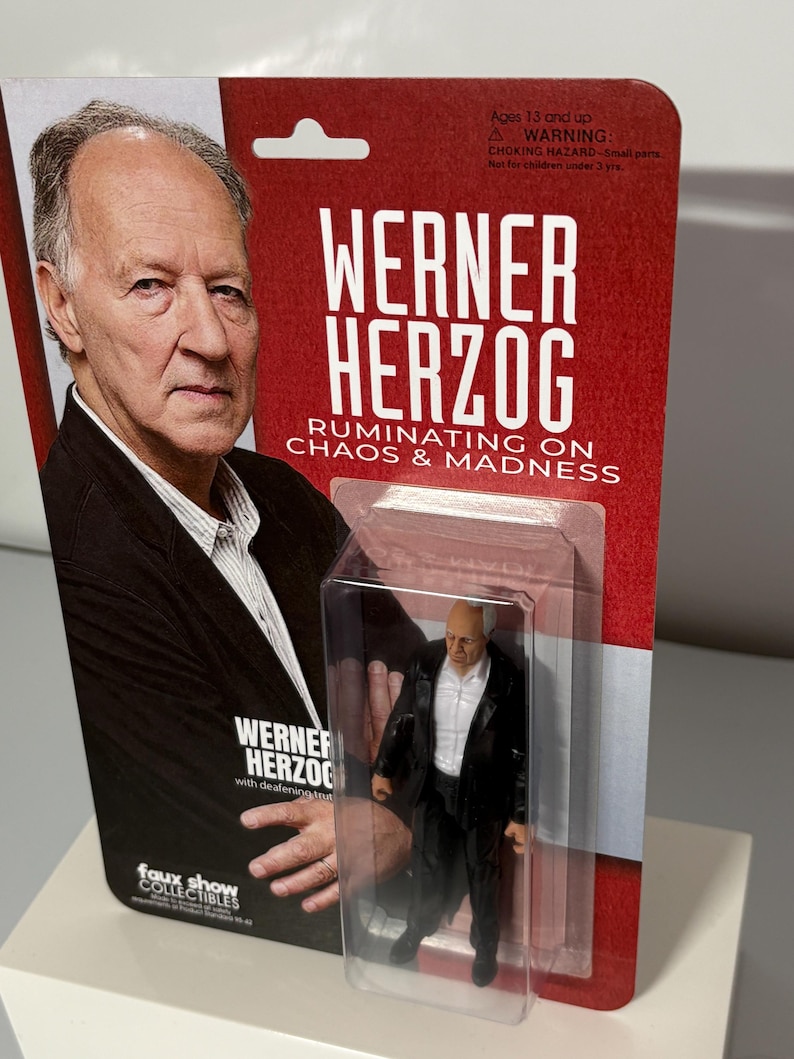 K&ouml;nnte beinhalten: Sammelfigur von Werner Herzog in einer transparenten Kunststoffverpackung. Die Verpackung zeigt ein Foto von Herzog und den Text "WERNER HERZOG RUMINATING ON CHAOS & MADNESS". Die Figur tr&auml;gt einen schwarzen Anzug und ein wei&szlig;es Hemd.