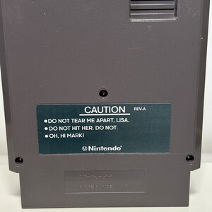 K&ouml;nnte beinhalten: Eine graue Nintendo Entertainment System (NES) Spielkassette mit einem schwarzen Etikett, auf dem "CAUTION" und "DO NOT TEAR ME APART, LISA. DO NOT HIT HER. DO NOT. OH, HI MARK!" steht. Das Etikett enth&auml;lt auch das Nintendo-Logo und den Text "REV-A". Die Unterseite der Kassette tr&auml;gt das Nintendo-Logo und den Text "PAT. PEND. MADE IN JAPAN."