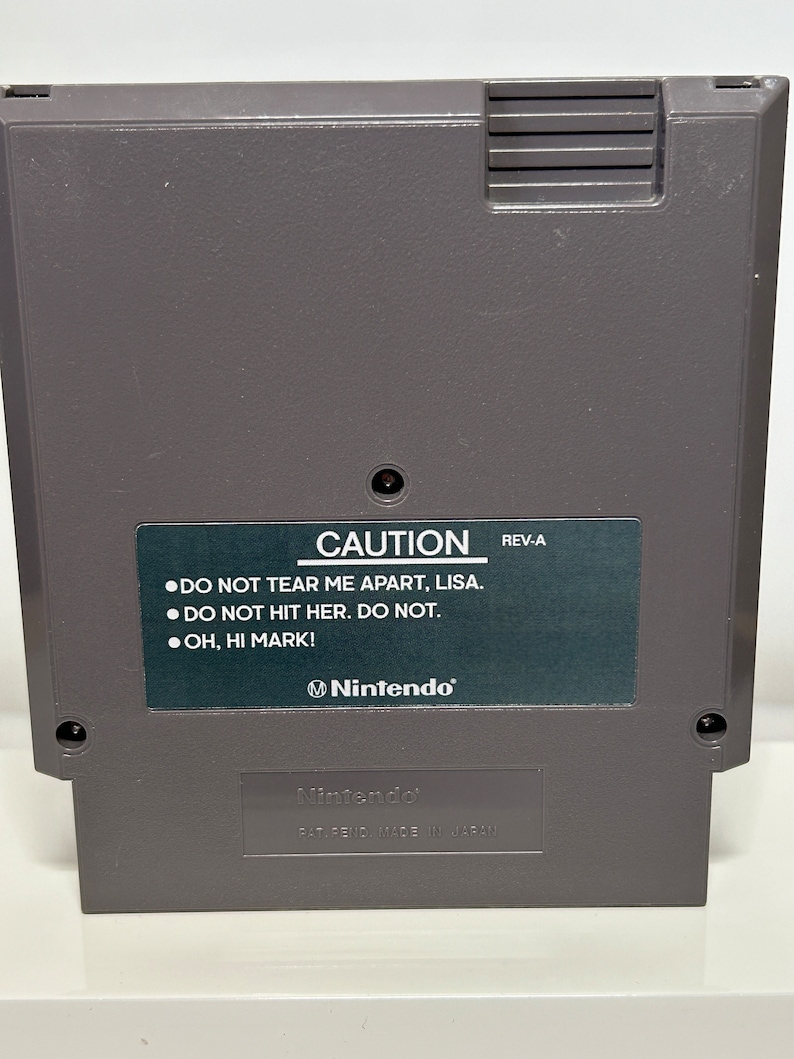K&ouml;nnte beinhalten: Eine graue Nintendo Entertainment System (NES) Spielkassette mit einem schwarzen Etikett, auf dem "CAUTION" und "DO NOT TEAR ME APART, LISA. DO NOT HIT HER. DO NOT. OH, HI MARK!" steht. Das Etikett enth&auml;lt auch das Nintendo-Logo und den Text "REV-A". Die Unterseite der Kassette tr&auml;gt das Nintendo-Logo und den Text "PAT. PEND. MADE IN JAPAN."