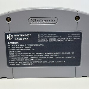 K&ouml;nnte beinhalten: Eine graue Nintendo 64-Spielkassette mit einem schwarzen Etikett. Das Etikett zeigt das Nintendo 64-Logo, die Worte "Game Pak" und eine Warnmeldung. Der Text auf dem Etikett enth&auml;lt Copyright-Informationen, Patentnummern und Anweisungen, um sich auf die Verbraucherinformationen und Vorsichtsma&szlig;nahmen im Booklet zu beziehen.