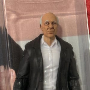 K&ouml;nnte beinhalten: Actionfigur eines &auml;lteren Mannes mit wei&szlig;em Haar, bekleidet mit einer schwarzen Jacke &uuml;ber einem wei&szlig;en Hemd mit Kragen. Die Figur befindet sich in einer transparenten Kunststoffverpackung mit rosa Hintergrund.